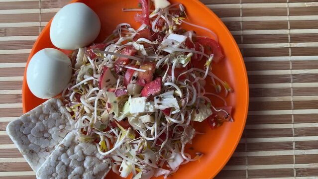 A fresh vegetable salad. Tomatoes, eggs, bread rolls, onions, apples, mound of sprouted mung beans in an orange bowl on bamboo background. Concept of diet, vegan, healthy products. Close up.