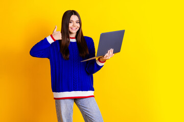 Photo portrait of pretty teen girl hold netbook thumb up wear trendy knitwear blue outfit isolated on yellow color background