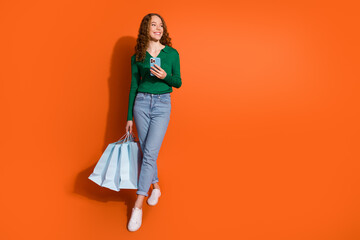 Photo of positive cheerful lady dressed green shirt holding shoppers texting device empty space isolated orange color background