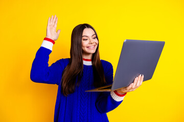 Photo portrait of pretty teen girl hold netbook video call wave hand wear trendy knitwear blue outfit isolated on yellow color background