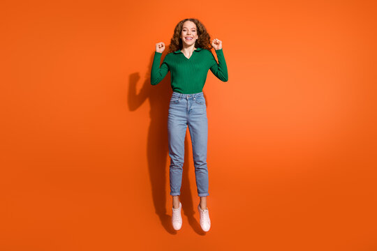Full length photo of lucky excited lady dressed green shirt jumping high rising fists isolated orange color background