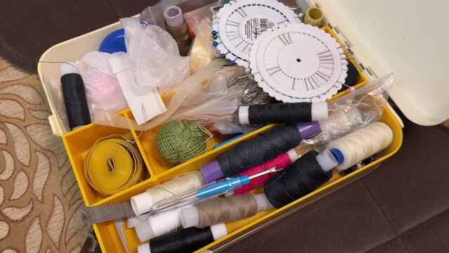 A lot of sewing items such as buttons, colorful fabrics, scissors, measuring tape, thimble, spools of thread on sewing pattern located in plastic box on the sofa. Close-up