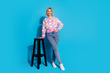 Full length photo of lovely girl wear checkered pullover pants lean on bar chair hold palm in pocket isolated on blue color background