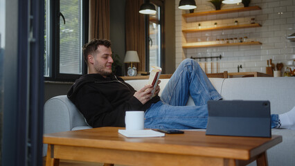 Carefree young man spends the weekend at home relaxing at home on the sofa with book read...