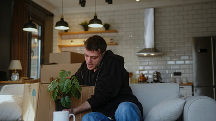 Young man moving into new apartment or moving out of house, sitting on sofa with carton boxes while...