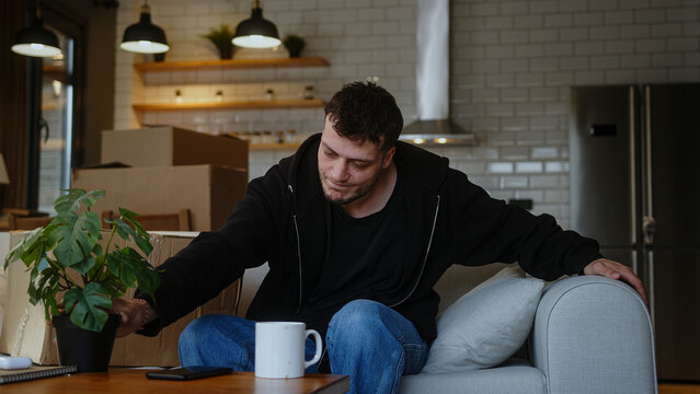 Young man feeling stressed moving into new apartment or moving out of house, sitting on sofa with carton boxes, tired of long renovation relocation processes