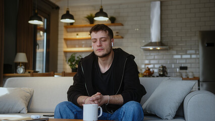 Young man sitting on sofa and listening to music with wireless in-ear headphones while drinking coffee or tea from mug. Man having fun, spending time alone at home
