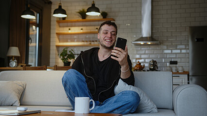 Smiling man sitting on sofa holding mobile phone, waving and greeting while having video conference call on smartphone. Man looking at screen, talking, smiling, enjoying online chat at home.