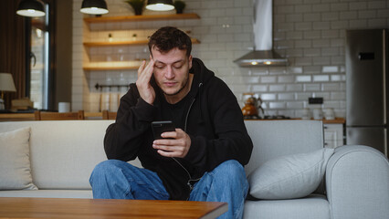 Young adult man sitting on sofa having headache while using smartphone. Tired young man get having...
