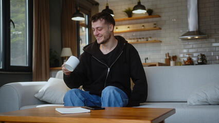 Funny guy sitting on the couch looking at the camera, talking, showing the mug, drinking coffee or...