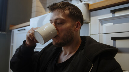 Man sitting on the kitchen floor fills a mug with water from a teapot and drinks.