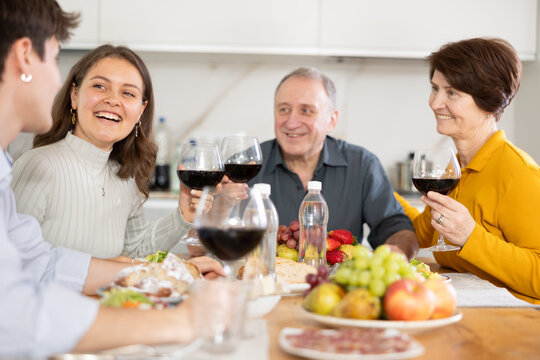 Smiling young woman engaging in lively conversation with husband and elderly parents-in-law at family table with wine and fruits in cozy home kitchen