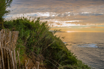 Fototapeta na wymiar Cliff trail with reed fence and wild vegetation with sunset on the horizon at sea in Lomba da Maia, São Miguel - Azores PORTUGAL