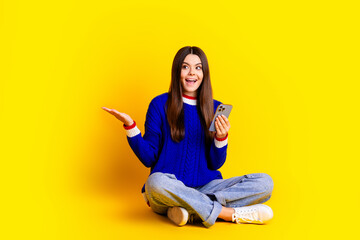 Full length photo of pretty teen girl hold device look empty space wear trendy knitwear blue outfit isolated on yellow color background