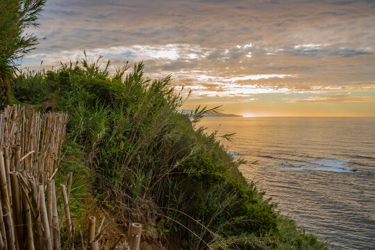 Cliff trail with reed fence and wild vegetation with sunset on the horizon at sea in Lomba da Maia, São Miguel - Azores PORTUGAL