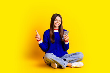 Full length photo of pretty teen girl hold device point empty space wear trendy knitwear blue outfit isolated on yellow color background