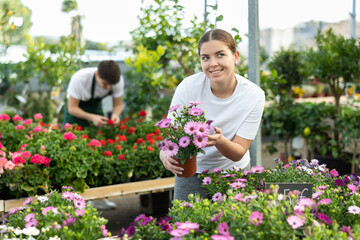 Smiling interested young girl choosing blooming potted African daisy with bright purple flowers in greenhouse to decorate country house in spring