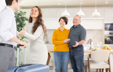 Happy young wife, beaming with smile, greeting husband back from trip with open arms while friendly elderly parents-in-law standing in background, reflecting warm family welcome