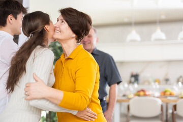 Fototapeta premium Positive young girl warmly welcoming with hug and kiss senior woman, coming family celebration at festive table with wine and light snacks