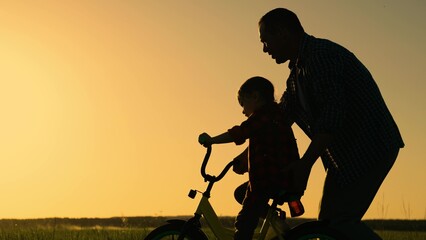 Dad teaches her little daughter to ride bicycle, sunset. Young father teaches child to keep balance while sitting on bicycle. Childhood dream to ride bike. Holiday family. Dad baby, Parental support.