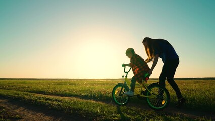 Active Mom teaches her little daughter to ride bike, sunset. Mother teaches her child to keep...