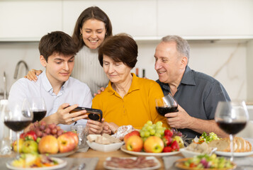 Cheerful lively multi-generational family gathered around set table at home dinner party in cozy kitchen, young adults sharing photos on smartphone with elderly parents