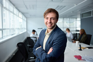 Happy young businessman in businesswear with arms crossed next to desk in office
