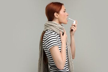 Young woman with scarf using throat spray on grey background - obrazy, fototapety, plakaty
