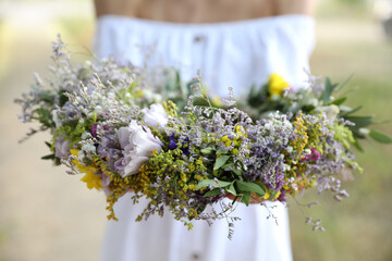 Young woman holding wreath made of beautiful flowers outdoors on sunny day, closeup