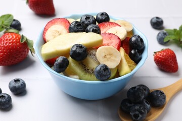 Tasty fruit salad in bowl and ingredients on white tiled table, closeup