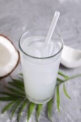 Glass of coconut water with ice cubes, palm leaf and nut on grey table