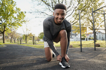 Happy young black man in sports clothing looking at camera while kneeling and tying shoelace in park - 784858842