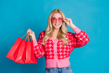 Portrait of adorable person dressed knitwear jumper touch pink sunglass holding new outfit in bags isolated on blue color background