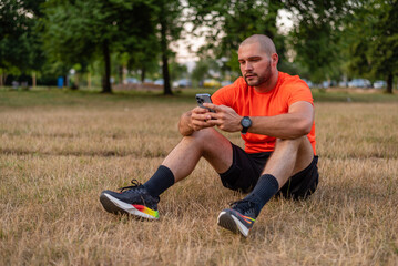 A young sporty man relaxing after exercise while using a phone in the park.