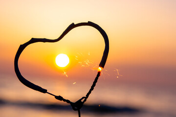 Heart-shaped sparkler burning against background of sea and rising sun at dawn. Bengal fire in...