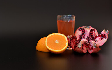 A glass of fresh fruit juice on a black background and pieces of orange and ripe pomegranate nearby.