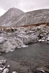 The stream of a small river flows through frozen ice formations surrounded by high snow-capped mountains on a cloudy autumn day.