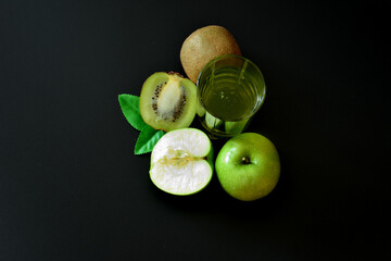 A glass of a mixture of fruit juices on a black background, next to pieces of a ripe kiwi fruit and half a green apple.