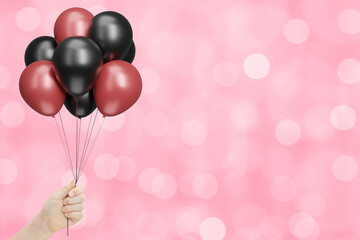 Female hand holds bunch of red and black balloons on blurred pink background. Empty space for text