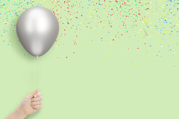 Female hand holds silver balloon on mint background with confetti. Empty space for text