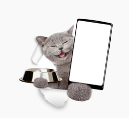 Happy cat looks through a hole in white paper, holds empty bowl and  shows smartphone with white blank screen in it paw, Empty free space for mock up, banner
