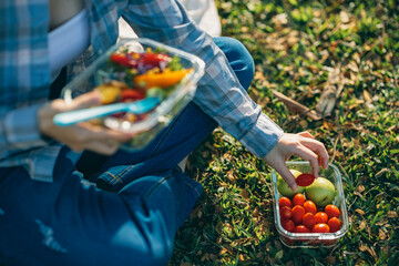 On a beautiful day, a woman enjoys a picnic in the park, The spread includes a glass lunch box...