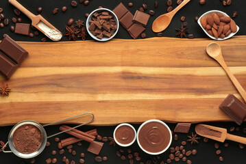 Wooden board with bowls of tasty melted chocolate, cocoa powder and almonds on black background