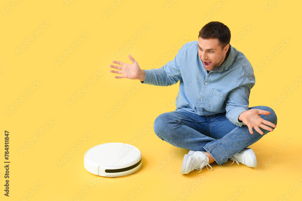 Sticker Surprised young man with robot vacuum cleaner sitting on yellow background