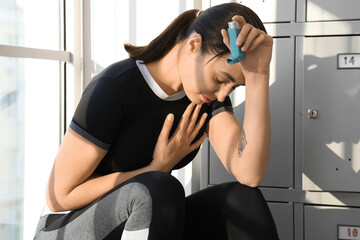 Sporty young woman with inhaler having asthma attack near locker in gym