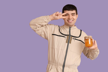 Male beekeeper with jar of honey showing victory gesture on lilac background