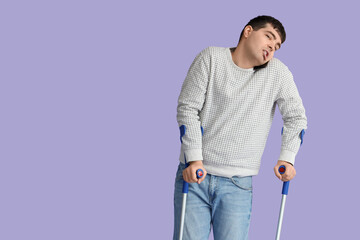 Young man with crutches talking by mobile phone on lilac background. National Cerebral Palsy...