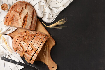Wooden board with sliced loaf of bread, wheat spikelets and grains on black background