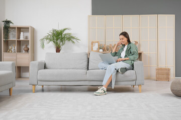 Pretty young woman with laptop sitting on grey sofa in living room