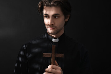 Young priest praying with cross on dark background, closeup